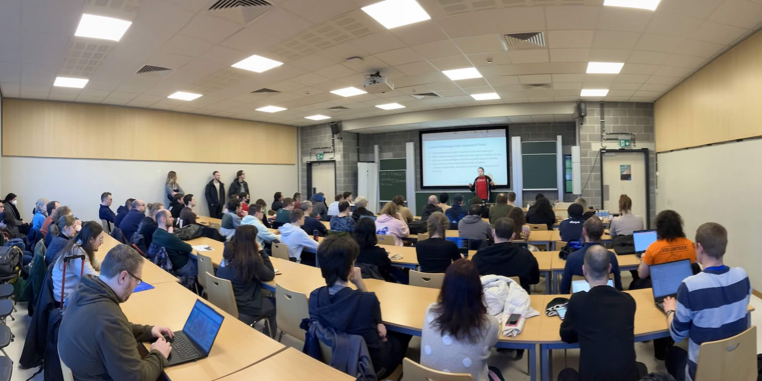 Panorama of the room during a talk, taken from the back. It’s a classroom with multiple rows of tables and seats. Some people are standing on the side. The speaker is in front of their slides at the front of the room.