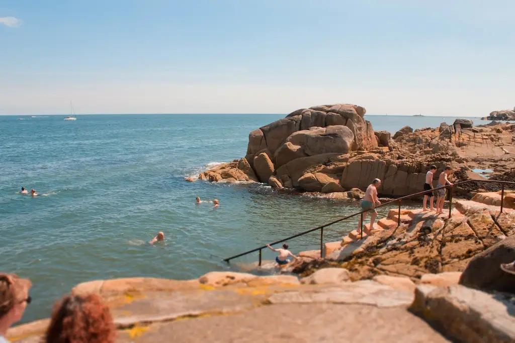 Photo of a sunny swimming spot on the Irish Sea, To the left is the sea with a boat in the distance and people swimming in the foreground. To the right are ocre and orange rock formations, with in the foreground a staircase going into the sea. There are people in swimsuits going up and down the stairs